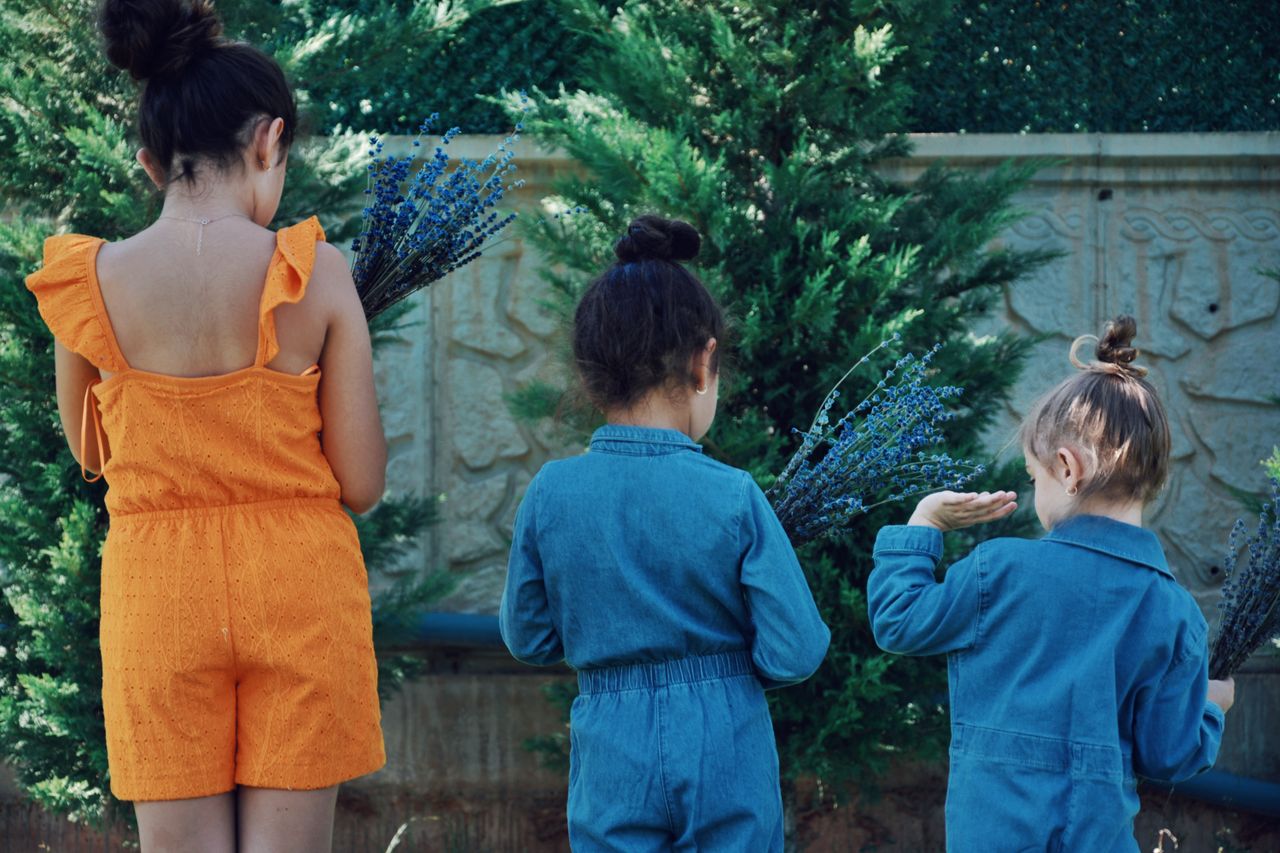 Three young sisters stand in an outdoor garden, holding lavender bouquets.