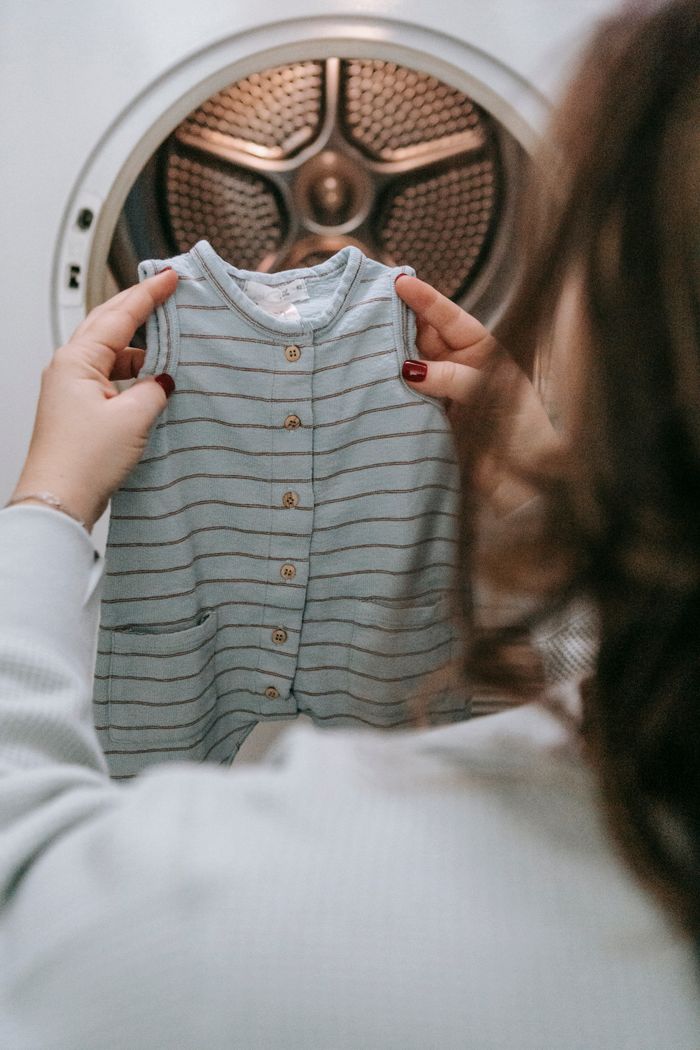 Back view of unrecognizable female with baby attire in hands near modern washing machine during laundry routine in light room
